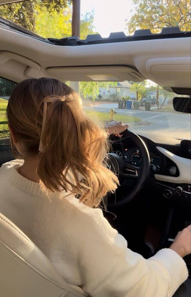 Person driving a car with sunroof open, sunlight illuminating hair and white sweater, picturesque neighborhood in view.