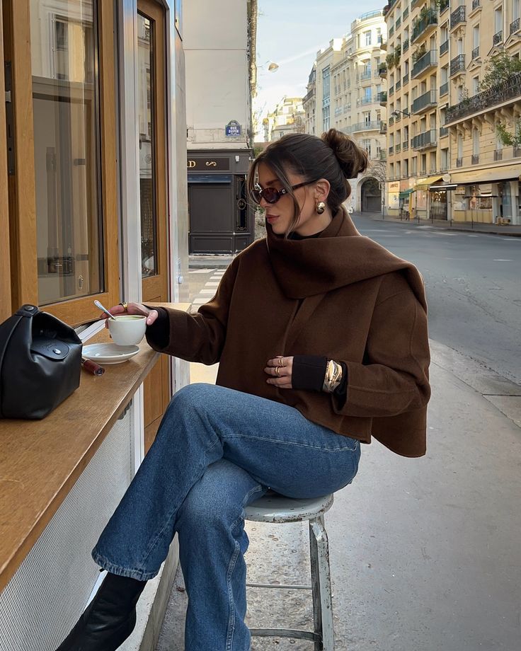 Woman enjoying coffee at an outdoor cafe, wearing a stylish brown coat and sunglasses, with a purse on the counter.