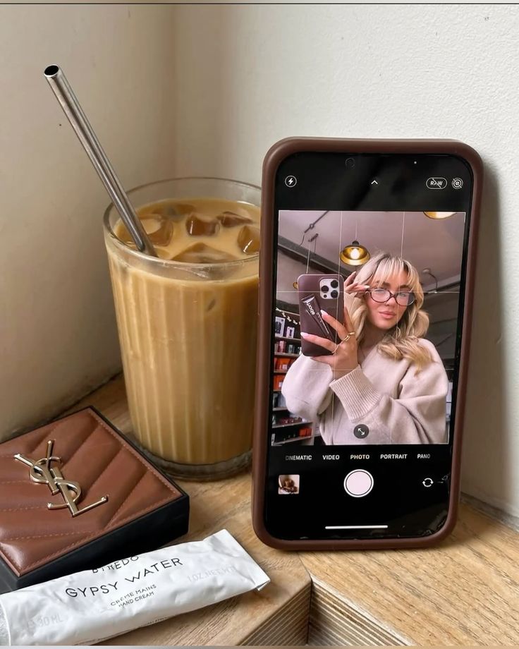 Smartphone showing a selfie next to iced coffee and luxury items on a wooden surface.