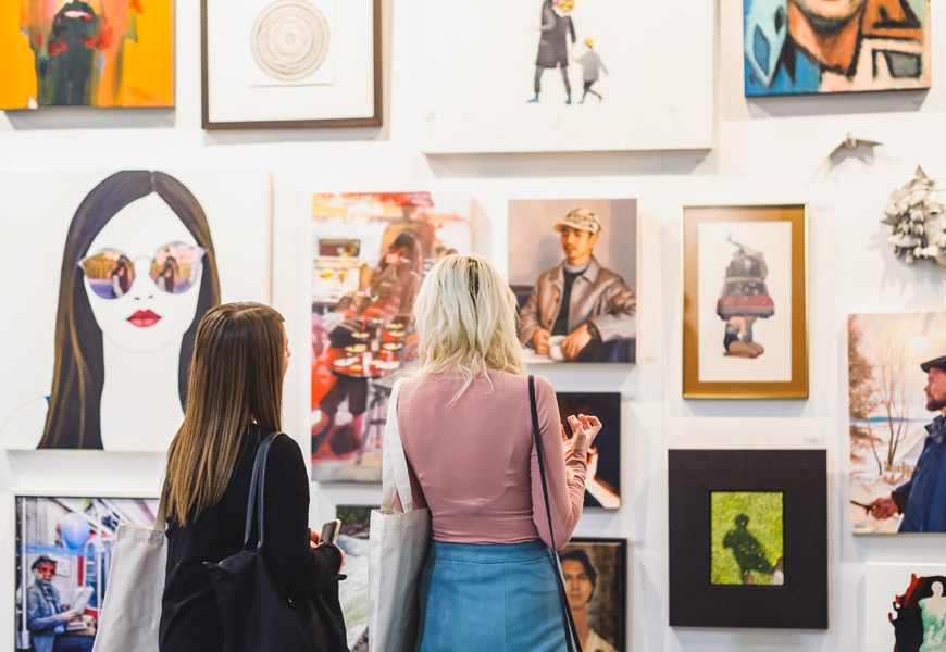 Two women viewing diverse art at a gallery, examining colorful paintings on a white wall.