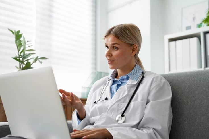 Doctor discussing patient care on video call with a laptop, wearing a stethoscope, sitting in a bright office.