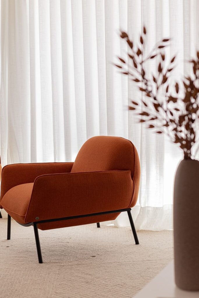 Modern orange armchair in a bright room with white curtains and decorative branches in a vase.