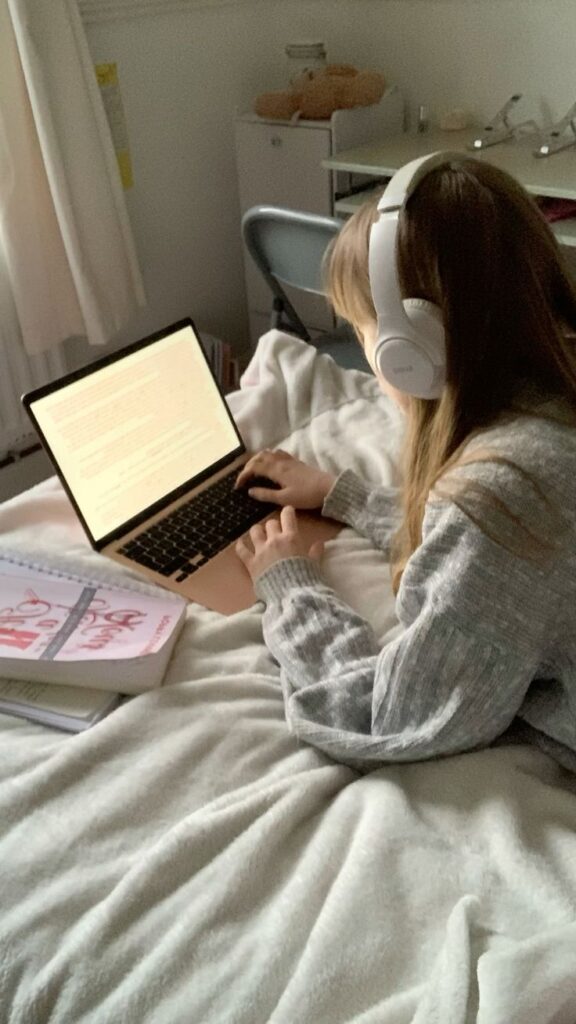 Person wearing headphones, working on a laptop in a cozy bedroom setting with notebooks around.