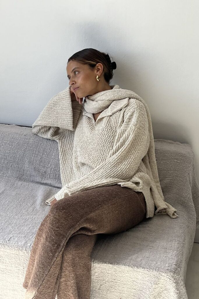 Woman in cozy beige sweater and scarf sitting thoughtfully on a gray couch.
