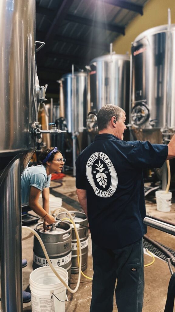 Crafting Excellence_ Aloha Beers Dedicated Brewers in Action Two workers managing beer kegs in a brewery with large fermentation tanks in the background. | Sky Rye Design Two workers managing beer kegs in a brewery with large fermentation tanks in the background.