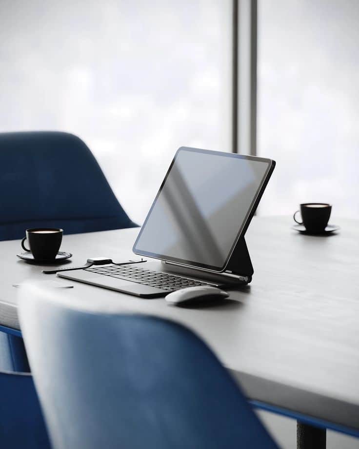 Creative Workspace Office _ VOZ Tablet with keyboard on a modern office table, surrounded by chairs and coffee cups, near large windows. | Sky Rye Design Tablet with keyboard on a modern office table, surrounded by chairs and coffee cups, near large windows.