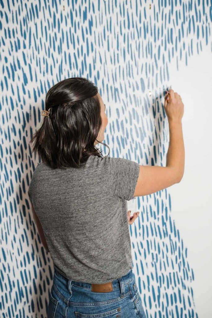 Woman painting blue abstract pattern on wall, wearing gray shirt and jeans.