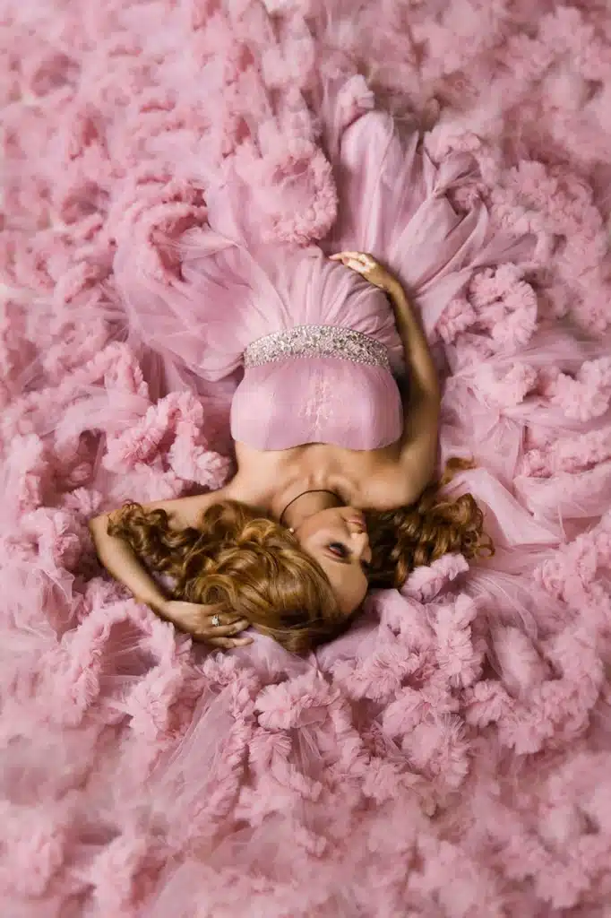 Woman in flowing pink gown lies on soft fabric, showcasing elegant fashion and serene beauty.