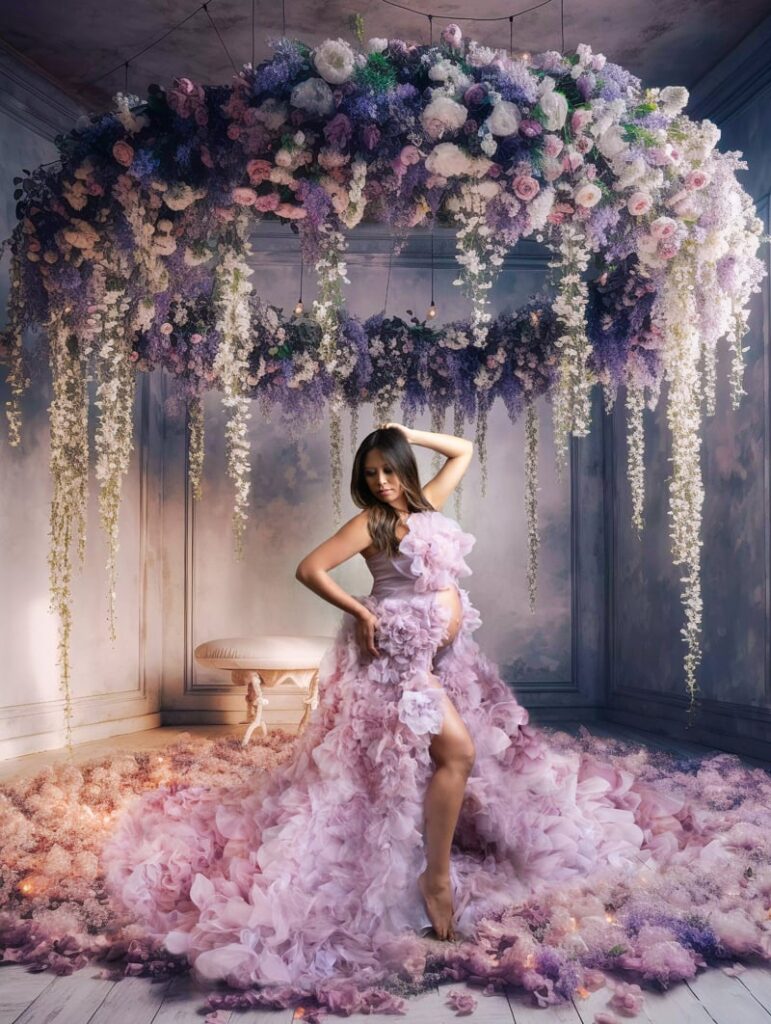 Pregnant woman in a floral pink gown poses under a vibrant flower arrangement, surrounded by petals.
