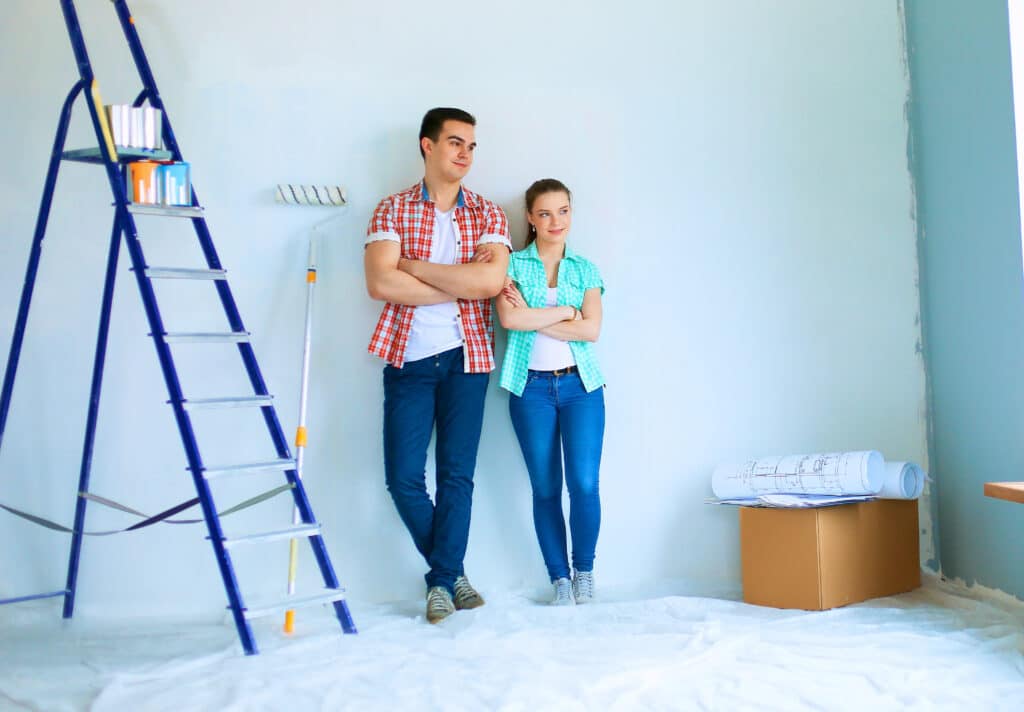Couple renovating room with ladder, paint roller, and blueprints, standing confidently beside each other.