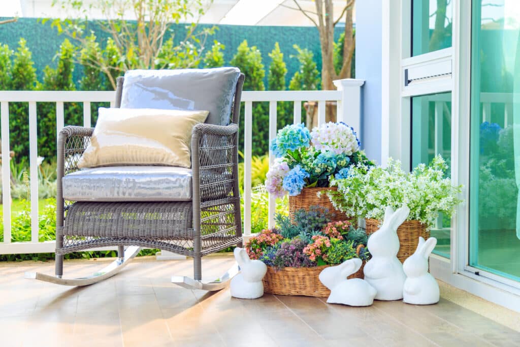 Cozy patio with a wicker rocking chair, floral baskets, and rabbit statues, ideal for relaxing in nature.