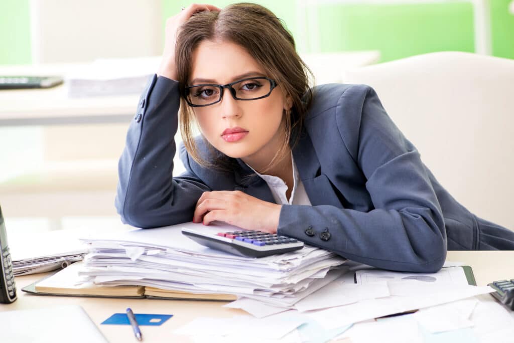 Businesswoman overwhelmed by paperwork, leaning on desk with calculator in office.