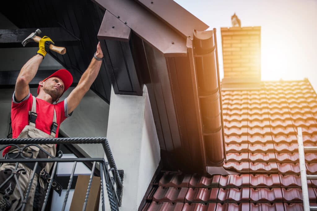Roofer in red uniform and cap repairing house roof under sunlight, holding hammer, inspecting wooden eaves.