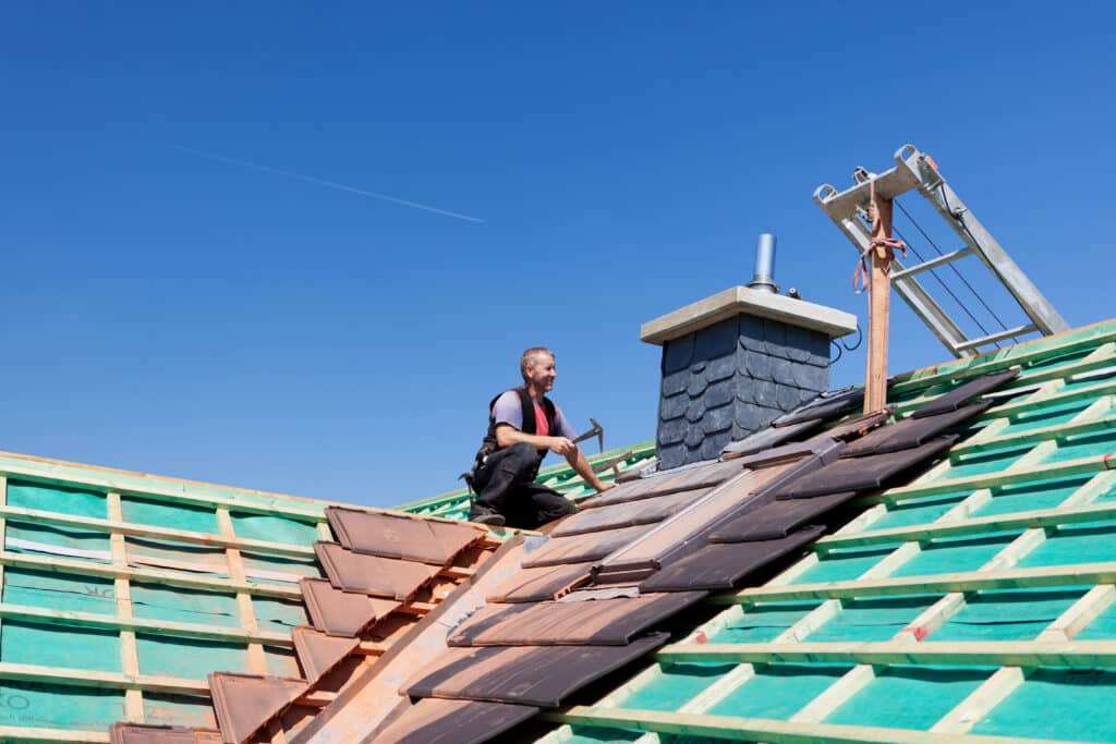 Roofer installing shingles on a house roof under clear blue sky, with chimney and ladder visible.