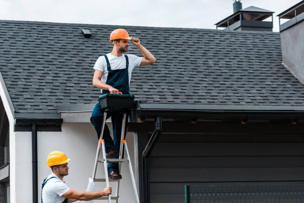 Two construction workers in helmets repairing a house roof, with one on a ladder.