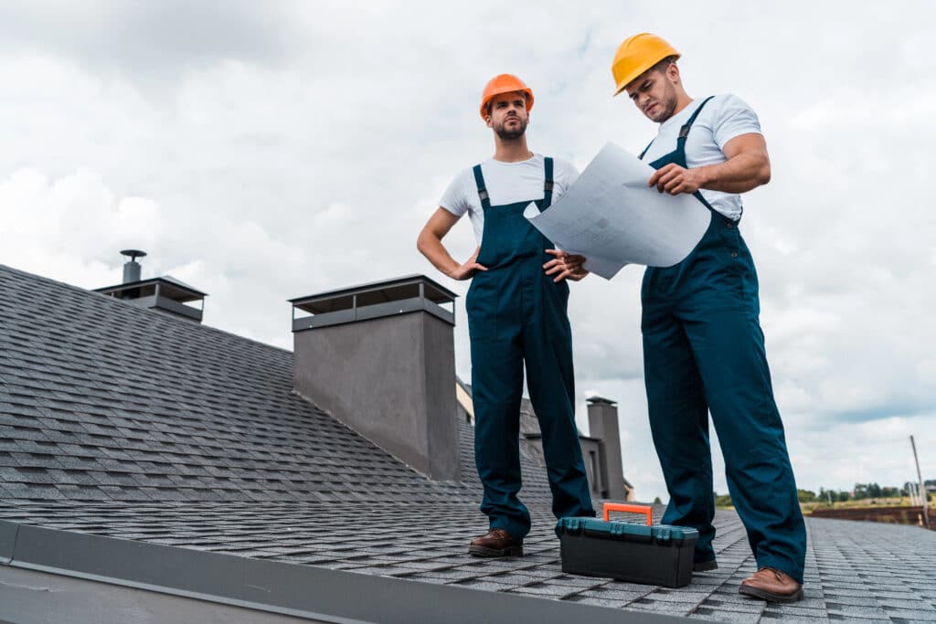 Two construction workers in helmets reviewing blueprints on a rooftop.