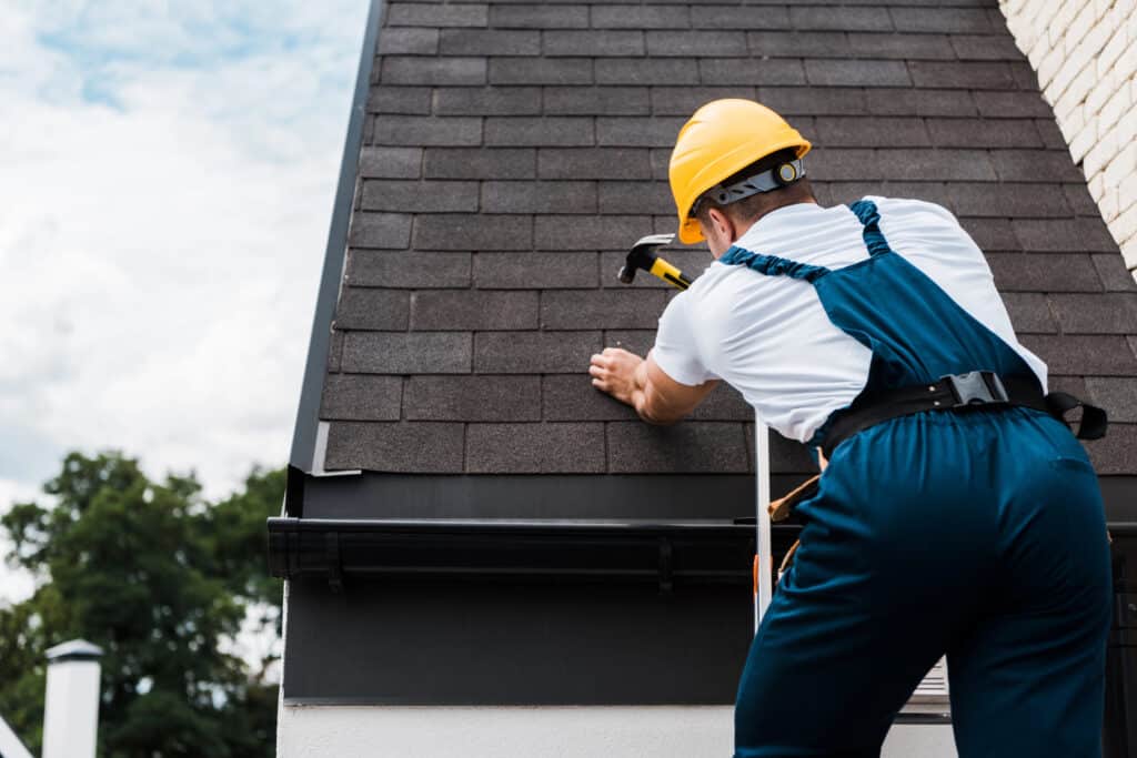 Roofer wearing a hard hat fixing shingles on a roof with a hammer on a clear day.
