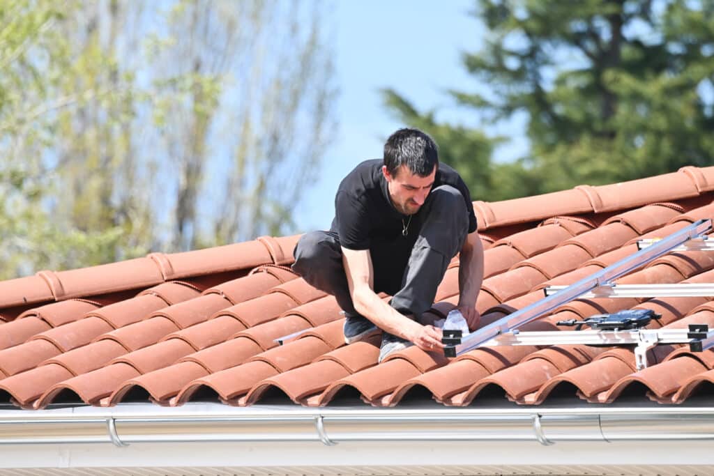 Worker installing solar panels on red tiled roof with green trees in background.