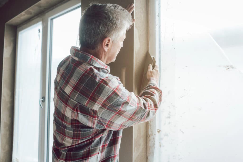 Man in plaid shirt applying plaster to window frame indoors.