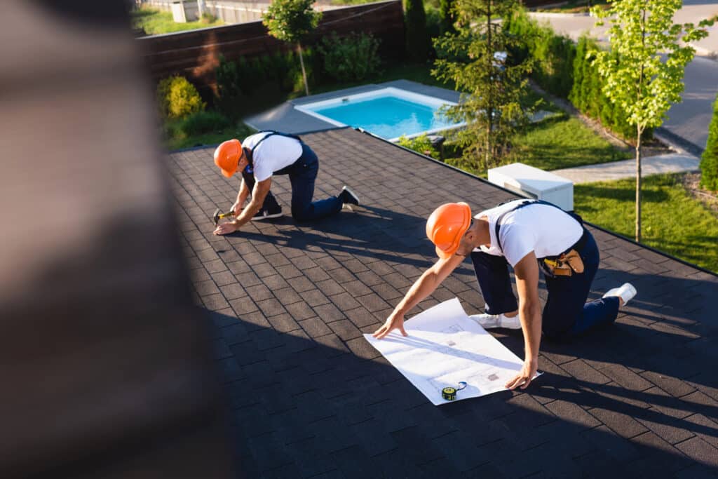 Roofers in orange helmets working on a roof with plans and tools, pool in the background.