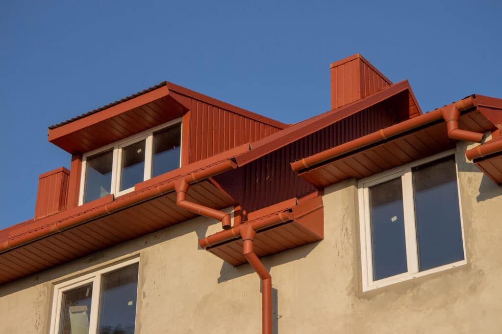 Modern house with red metal roofing, dormer windows, and gutters under a clear blue sky.