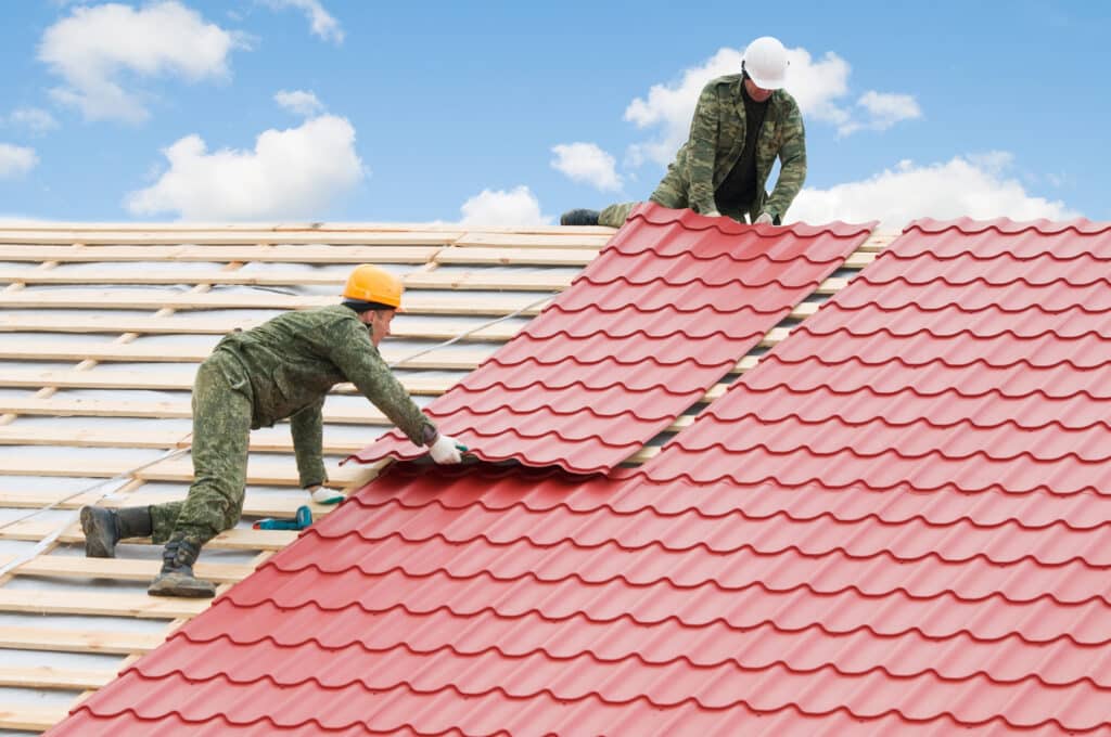 Roofers installing red metal sheets on a roof under blue sky.