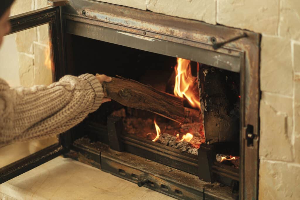 Depositphotos_616877178_XL Person placing a log in a cozy fireplace, enhancing warmth and ambiance in a living room during winter. | Sky Rye Design Person placing a log in a cozy fireplace, enhancing warmth and ambiance in a living room during winter.
