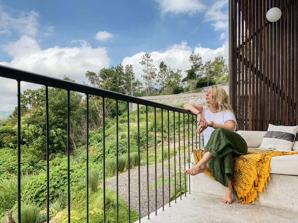 Woman relaxing on a balcony surrounded by greenery, enjoying a serene view under a blue sky.