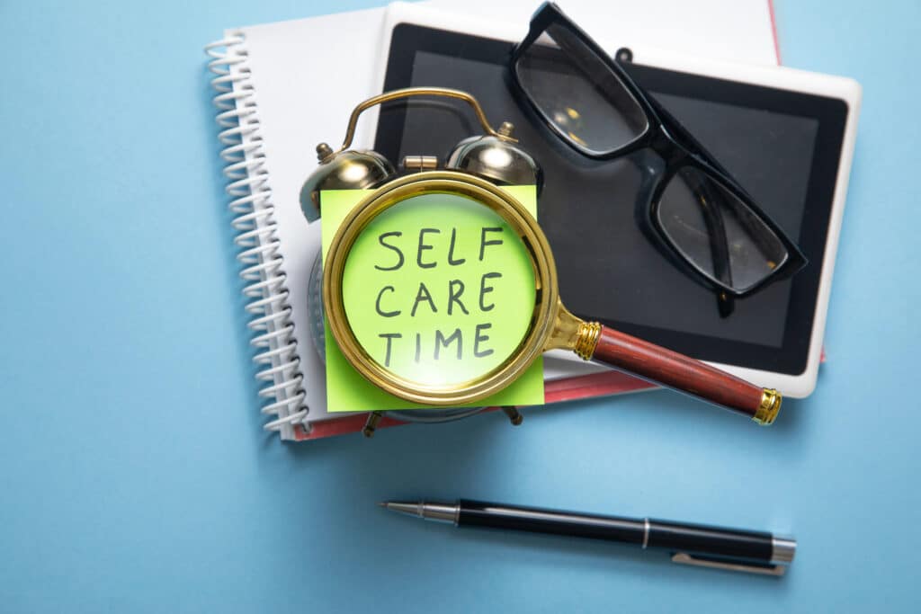 Magnifying glass over Self Care Time note, near tablet, glasses, pen, and notebooks on blue background.
