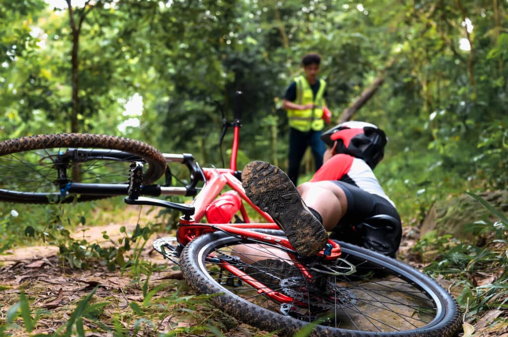 Cyclist on forest trail after fall, wearing helmet, with bike on ground, helper approaching.