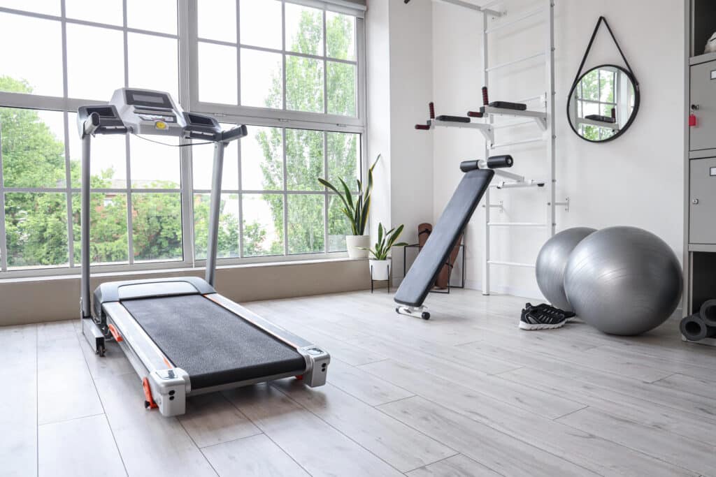 Modern home gym with treadmill, incline bench, yoga balls, and light wood flooring, with large windows and plants.
