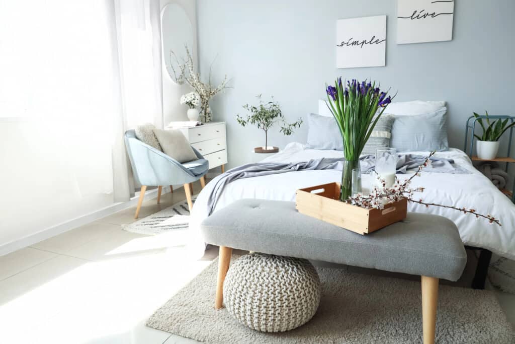 Modern bedroom with minimalist decor, featuring a gray bed, cozy chair, and fresh flowers on a bench.