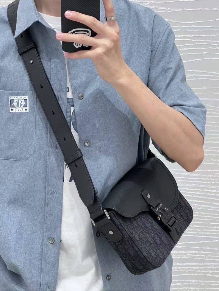 Man in blue shirt with smartphone, wearing a stylish black crossbody bag, capturing a mirror selfie.