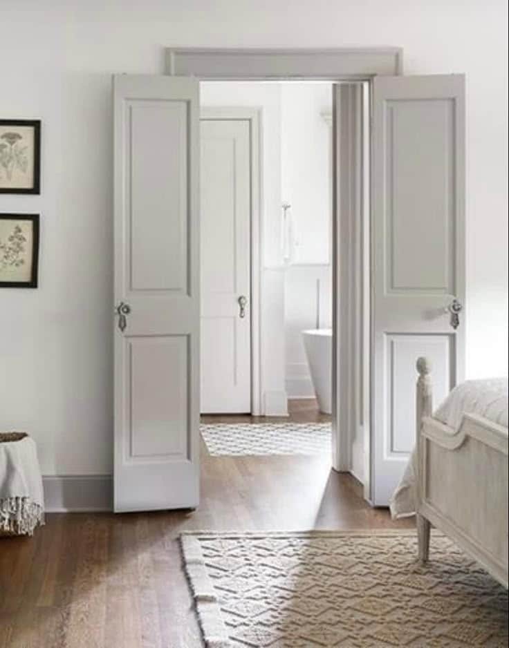 Open white double doors leading to bathroom in a bright, minimalist bedroom with wooden floors and cozy rugs.