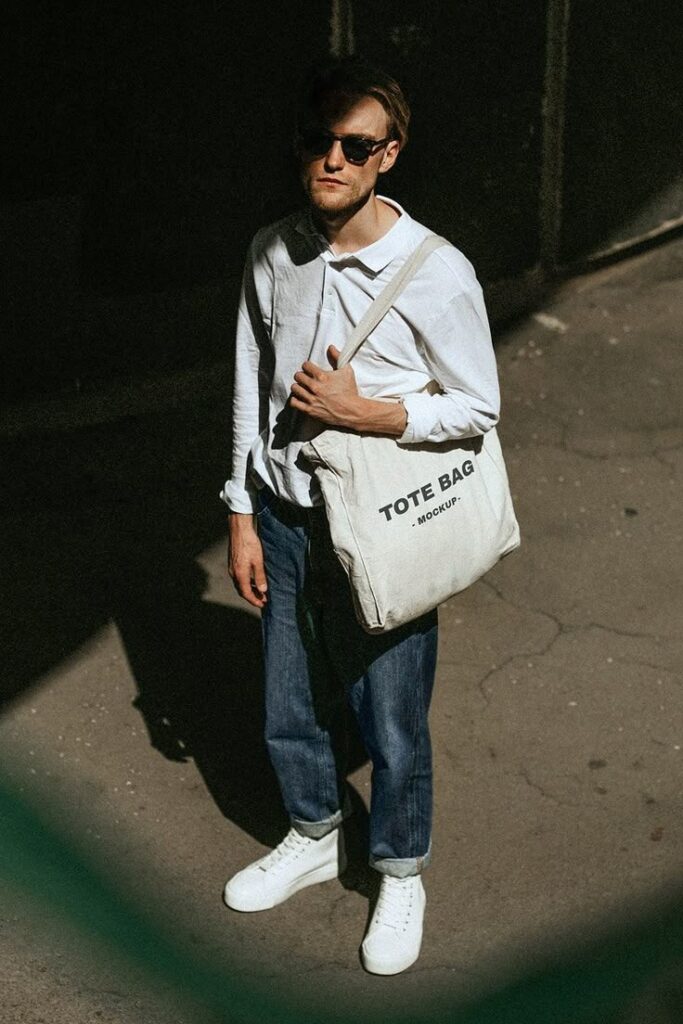 Man in casual attire with sunglasses and a white tote bag, standing on a sunlit street.