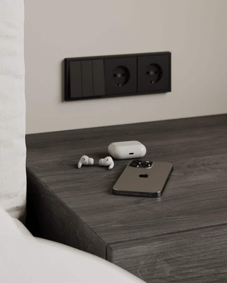Smartphone and wireless earbuds on a dark wood bedside table with dual power outlets in modern bedroom setting.