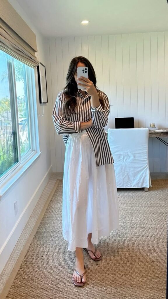 Woman in striped shirt and white skirt taking mirror selfie at home.