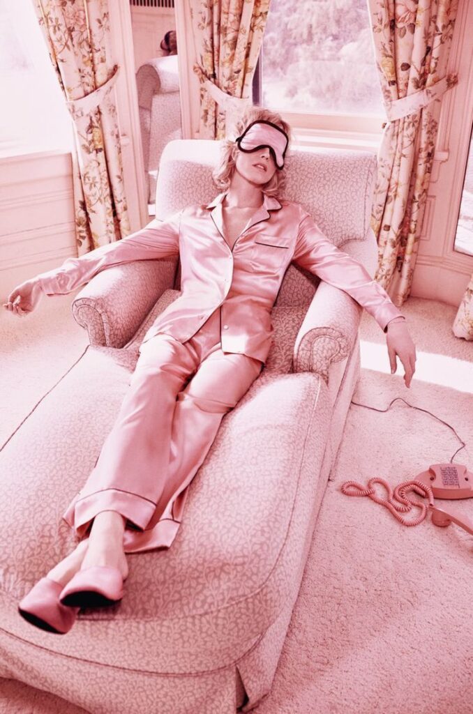 Person lounging in pink satin pajamas with sleep mask, relaxing on a plush chair by a window with floral curtains.