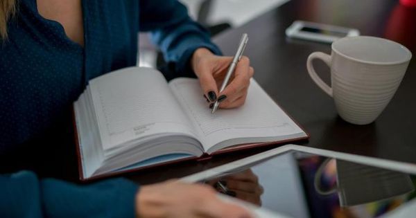 Person writing in a notebook at a desk with a cup and tablet, focusing on work or study tasks.