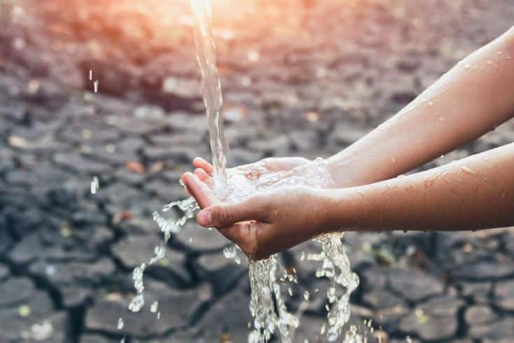 Hands catching flowing water above dry, cracked earth under sunlight, symbolizing hope and scarcity.