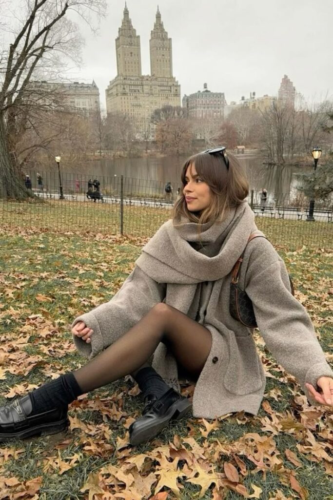 Woman in winter coat relaxing on Central Park lawn with NYC skyline in background.