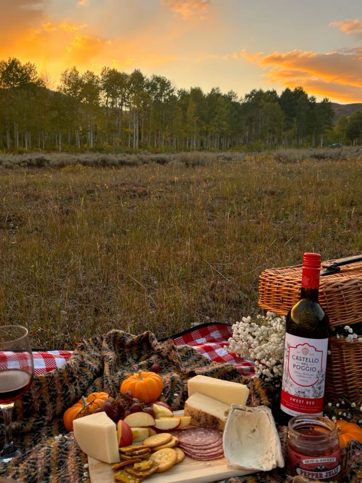 Sunset picnic with wine, cheese, and snacks on a blanket in a picturesque autumn field, surrounded by trees.