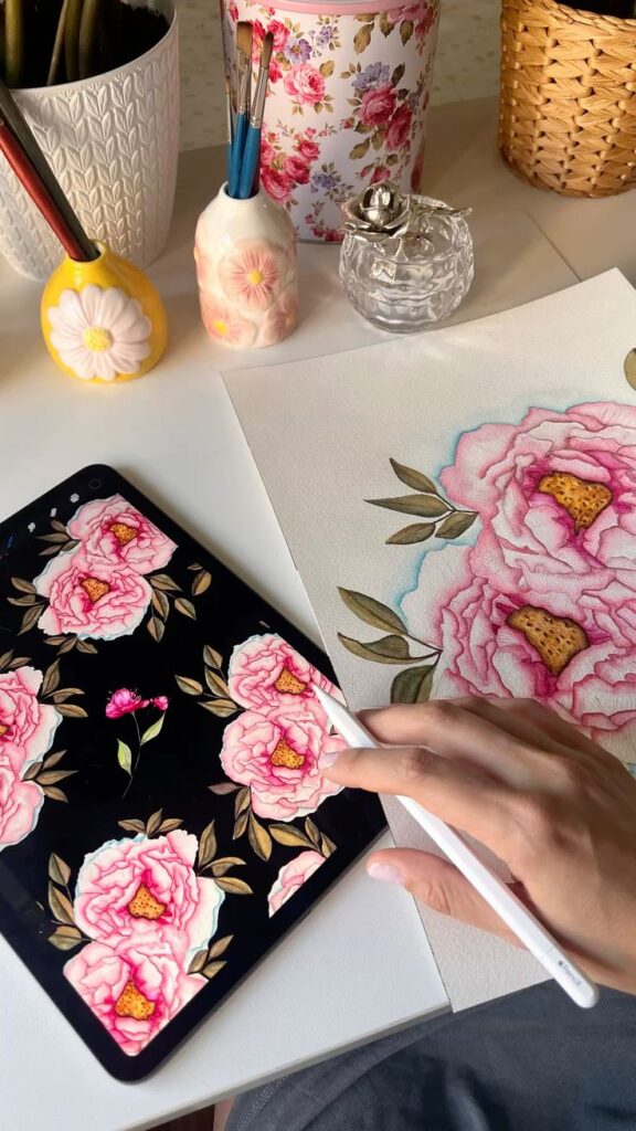 Artist drawing pink peonies digitally and on paper with brushes in floral holders nearby.