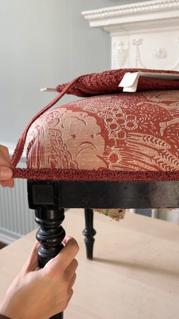 Hands adjusting fabric on a vintage wooden stool with a textured red pattern.