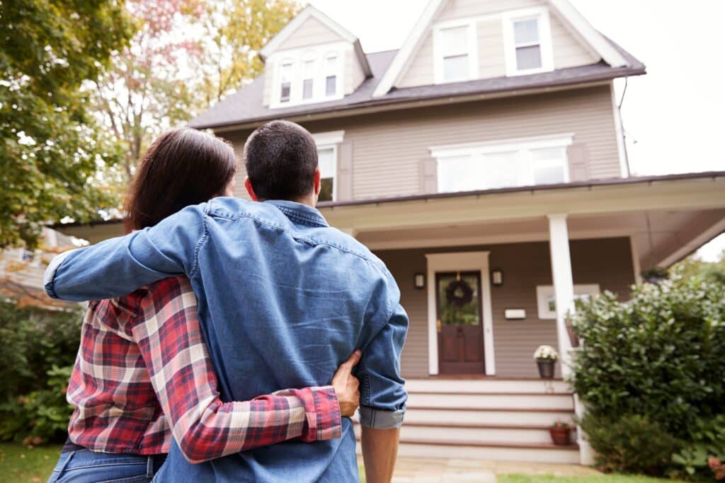 Couple embracing and looking at their new home with a porch, surrounded by trees and greenery.