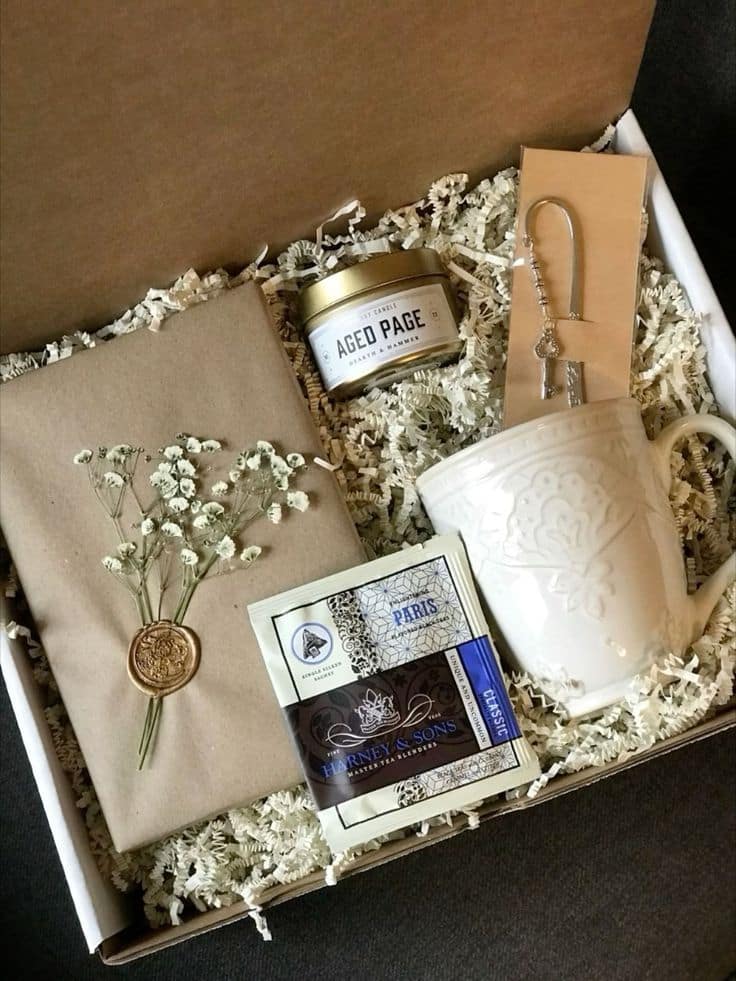 Gift box with a white mug, candle, tea, and a letter with flowers, arranged in decorative paper filling.