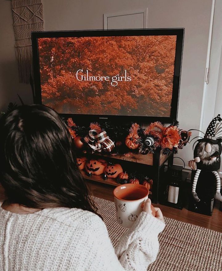 Cozy fall scene with person watching TV show Gilmore Girls, holding a pumpkin mug, surrounded by autumn decor.