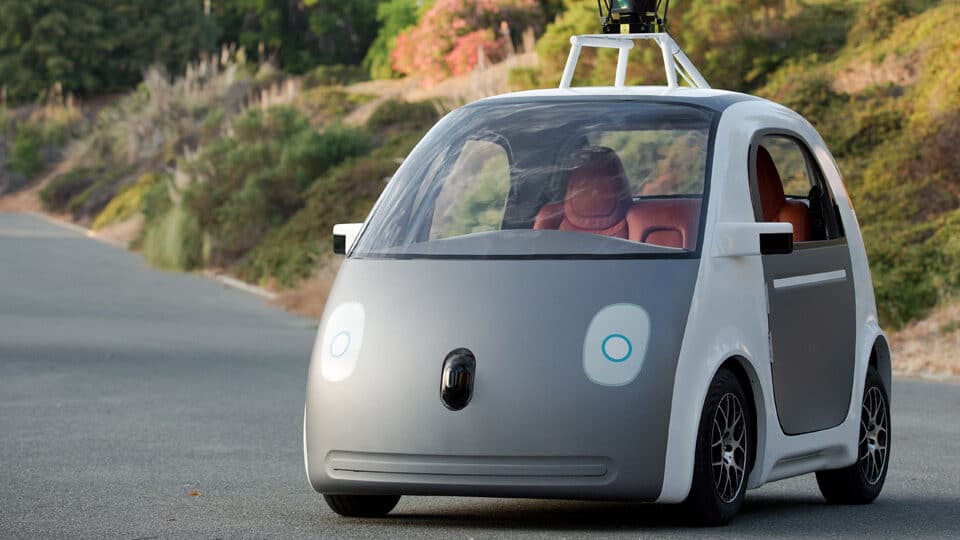 Autonomous white car driving on a scenic road lined with green trees and shrubs.