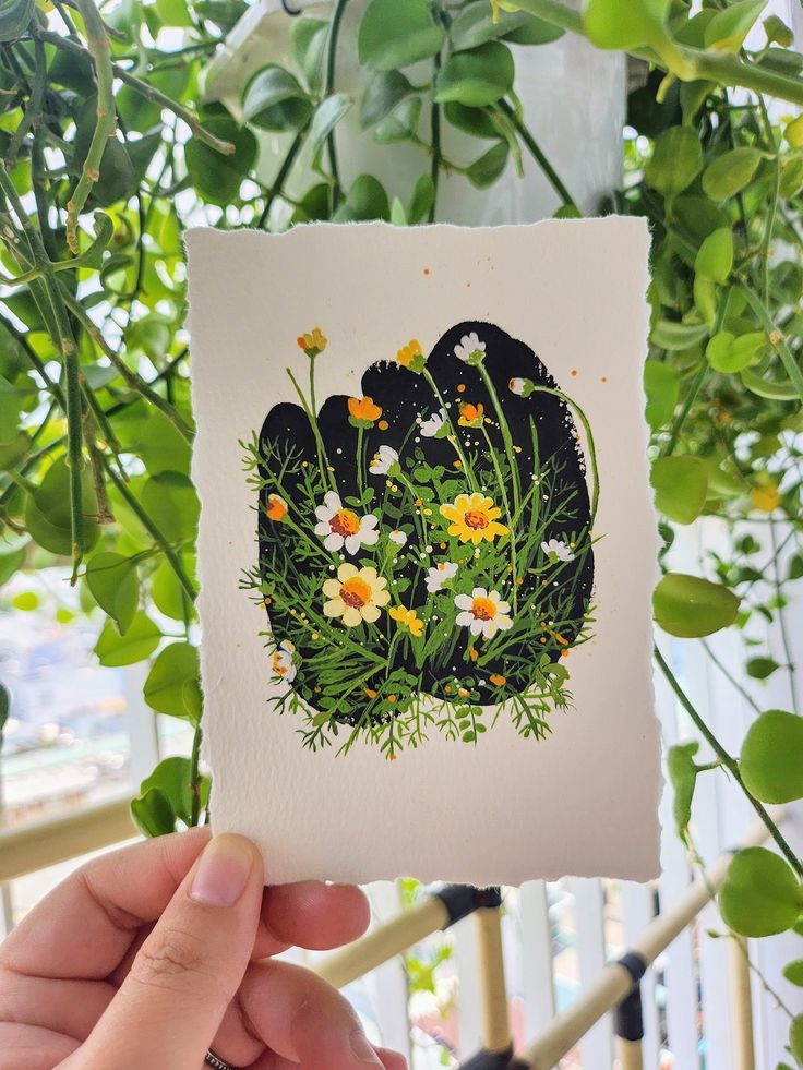 Hand holding a floral illustration of wildflowers, surrounded by lush greenery in a natural setting.