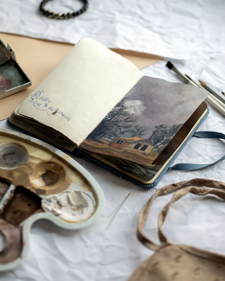 Open sketchbook with artwork beside a paint palette and brushes on a desk.
